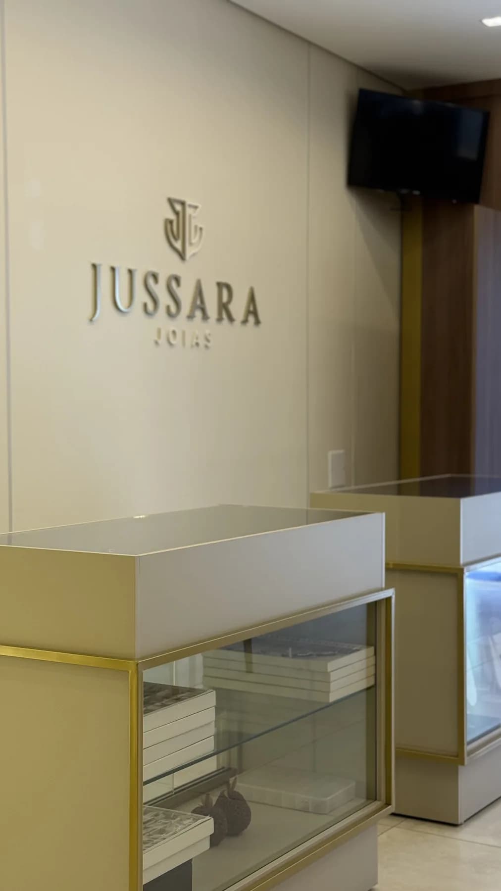 Parede da loja com o letreiro JUSSARA JOIAS em relevo dourado e, à frente, dois balcões de vitrine com estrutura de latão e bandejas de joias